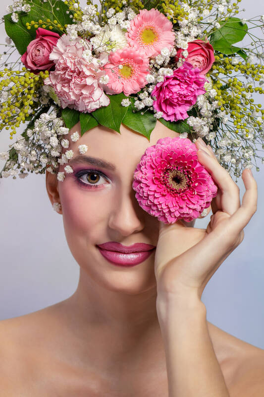 Portrait einer Frau mit Blumen-Kopfschmuck. mit der linken Hand hält sie sich die Blüte einer pinkfarbigen Gerbera vor das linke Auge.