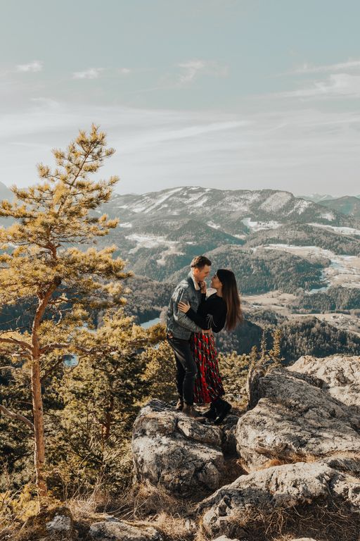 Ein Paar umarmt sich auf einem frühlingshaft bewaldeten Felsen mit Blick auf verschneite Berge. verschneite 
