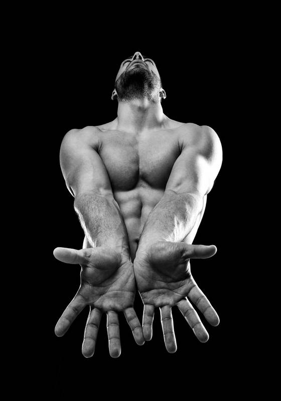 Ein muskulöser Mann mit unbekleidetem Oberkörper streckt die geöffneten Hände zur Kamera und sieht nach oben. Schwarz-Weiß.