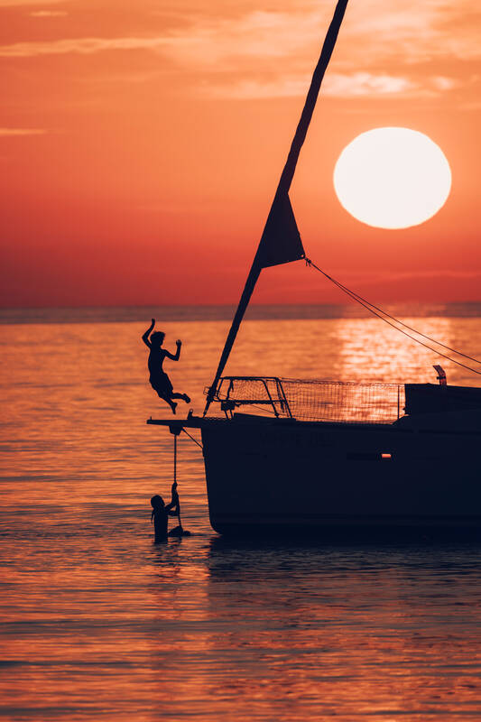 Ein Segelboot liegt im Sonnenuntergang vor Anker und Kinder springen vom Bug ins Wasser.