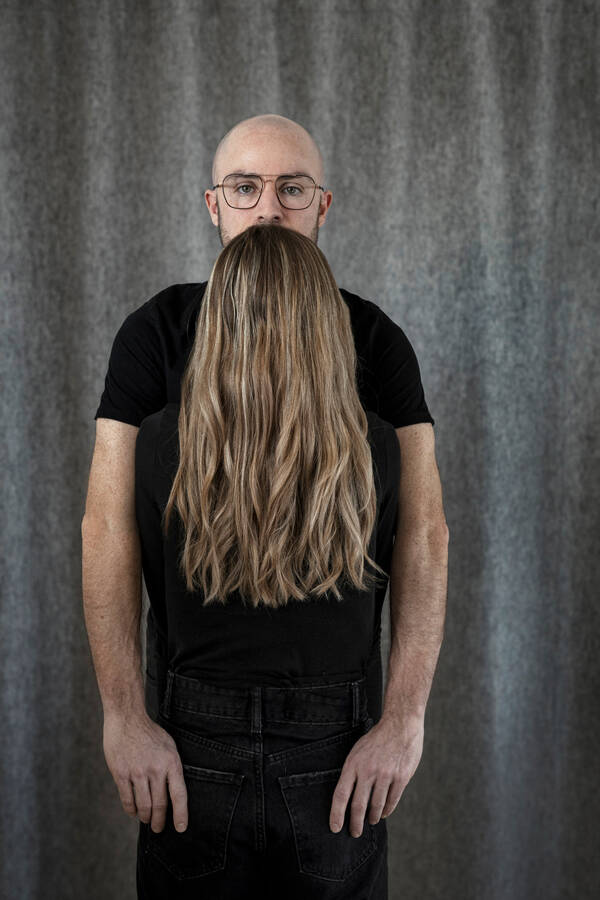 ein Mann mit Glatze und eine Frau mit langen Haaren, beide schwarz gekleidet, stehen so voreinander, dass auf den ersten Blick ihre Haare wie sein Bart wirken.