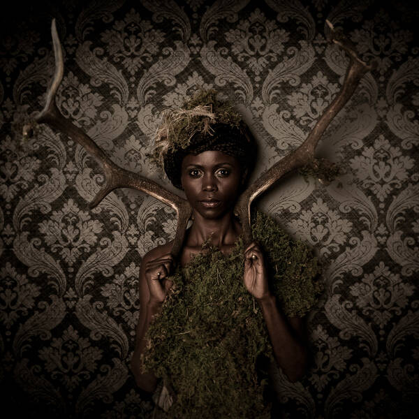 Eine dunkelhäutige Frau in Moos gehüllt steht mit zwei Hirsch-Abwurfstangen auf der Schulter vor einer gemusterten Tapete.