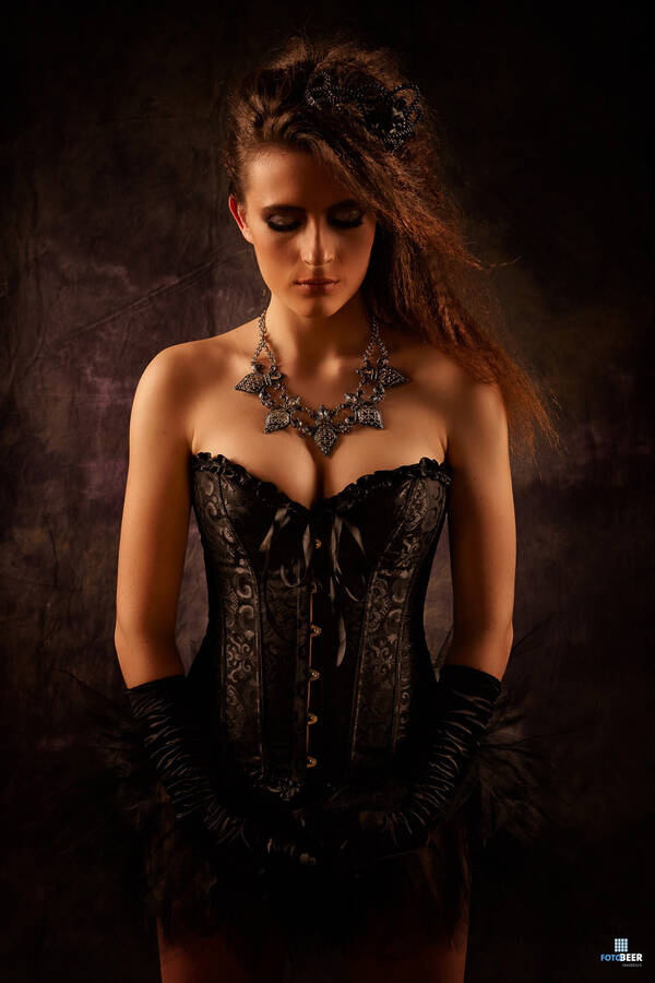 Eine junge langhaarige Frau in schwarzer Korsage und Handschuhen steht mit geschlossenen Augen vor einer dunklen Wand.