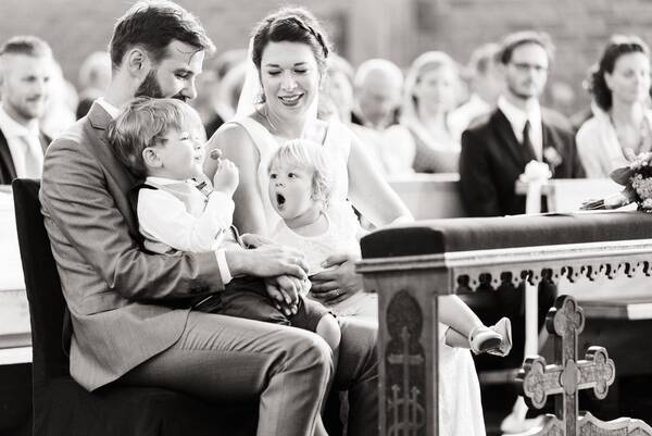 Ein Brautpaar in der Kirche sitzend, zwei Kinder am Schoß, eines hat einen Lolly, das andere schaut mit geöffnetem Mund auf den Lolly. Schwarz-Weiß.
