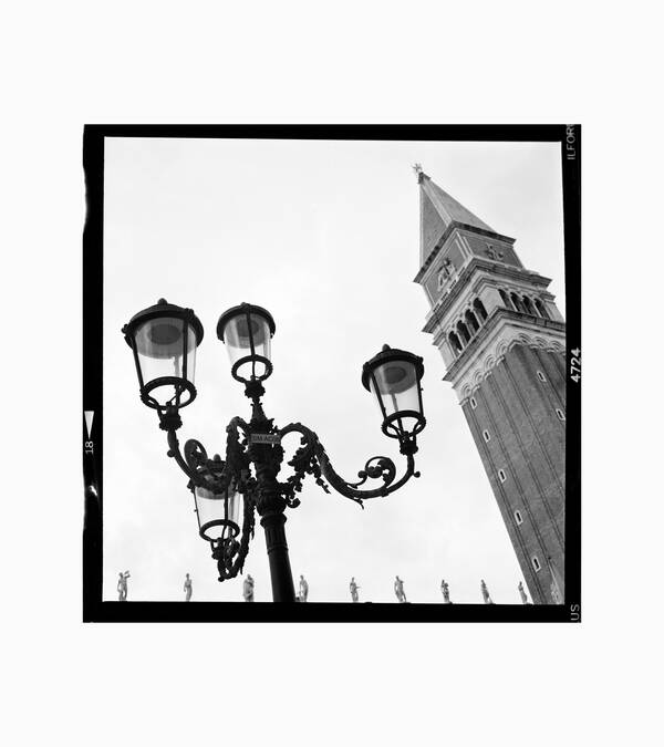Der Campanile von San Marco, im Vordergrund eine Straßenlaterne, am unteren Bildrand sieht man eine reihe Marmorfiguren. Schwarz-Weiß.