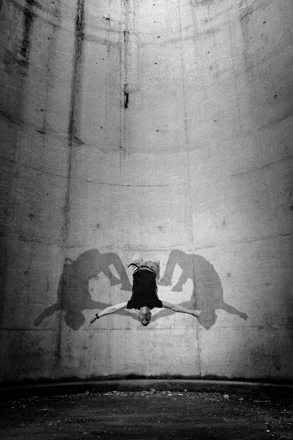 Ein Mann im Sprung schwebt scheinbar schwerelos kopfüber in einem Betonsilo. Blitzlichter zeichnen symetrisch seinen Schatten links und rechts an die gekrümmte Rückwand. Schwarz-Weiß.