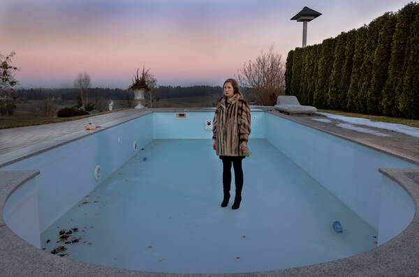 Eine Frau in Pelzjacke mit einer halbvollen Flasche in der Hand steht mit leerem Gesichtsausdruck in einem leeren Pool im winterlichen Abendlicht. Am Beckenrand steht ein Sektglas und eine nicht ganz leere Sektflasche liegt daneben.