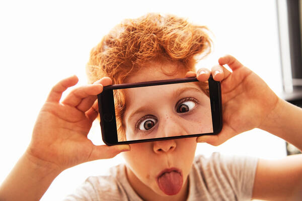 Ein rothaariges Kind hält sich ein Mobiltelefon vor das Gesicht auf dessen Display verdrehte Augen zu sehen sind, die gut zu der herausgestreckten Zunge des Kindes passen.