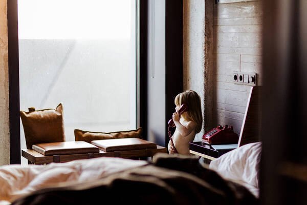 Ein Kind steht telefonierend neben einem Bett in einem Hotelzimmer und sieht aus dem Fenster