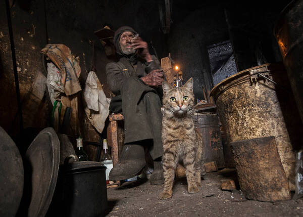 Ein Mann in schmutzigen Händen und Kleidern sitzt in einer Niesche und raucht, vor ihm sitzt eine Katze, rundherum alte Eimer und Töpfe.