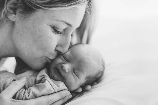 Nahaufnahme eines Neugeborenen, das von der Mutter in beiden Händen gehalten und auf die Wange geküsst wird. Schwarz-Weiß