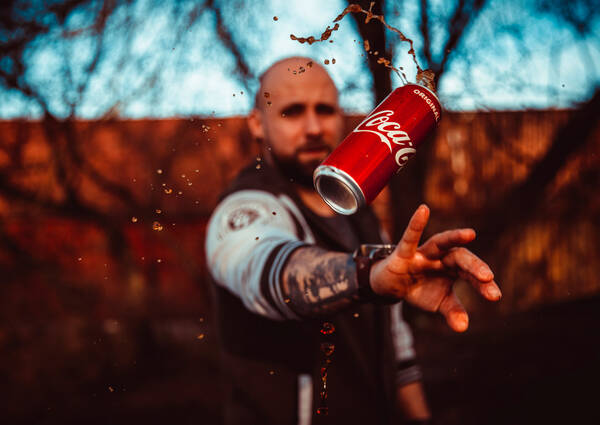 Eine Cola-Dose fliegt drehend in der Luft, unscharf im Hintergrund der Mann, der sie geworfen hat mit Bart und tätowiertem Unterarm.