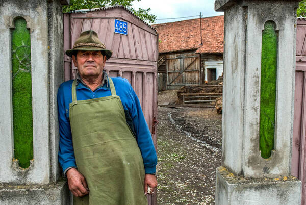 Bild eines alten Bauern mit grüner Schürze, der sich an eine Säule im Eingang zum Bauernhof lehnt.