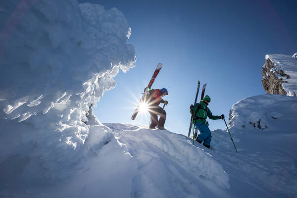 Zwei Skitourengeher haben die Schi auf die Rucksäcke geschnallt und gehen bei strahlend sonnigem Wetter über einen Graz neben einer Wächte.