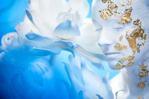 Portrait einer völlig weiß geschminkten und partiell mit Goldfolie beklebten Frau. im Hintergrund eine Blüte in weiß und blau.