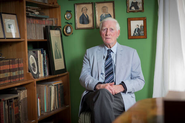 Portrait eines alten Mannes in hellblauem Sakko mit gestreifter Krawatte vor einer grünen Wand mit historischen Portraits und neben einer hölzernen Bücherwand in der auch Portraitbilder aufgestellt sind.