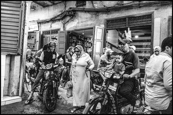 Turbulente Straßenszene in einer engen Gasse mit einem Durcheinander von Fußgängern und Motorrädern. Kleine Kinder sitzen auf dem Schoß der Motorradfahrer, eine alte Frauen mit Kopftuch trägt ihre Eikäufe. Schwarz-Weiß.