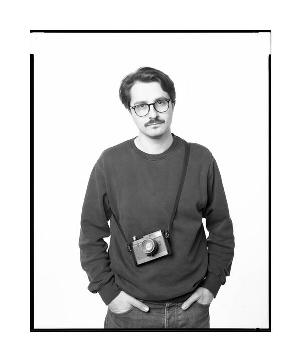 Portrait eines jungen Mannes mit Schnauzbart und Brille. Er steht mit den Händen in den Hosentaschen und eine Leica Kamera umgehängt. Schwarz-Weiß