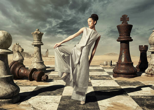 Eine june Frau in silbrig-weiß gewirkter Tunika steht in einer Pose die an Kampfsport erinnert auf einem überdimensionalen Schachfeld mit lebensgroßen Figuren in einer dystopischen Wünstenlandschaft. Während der weiße und der schwarze König noch am Spielfeld stehen, liegt die schwarze Dame so als wäre sie von der Frau, der 