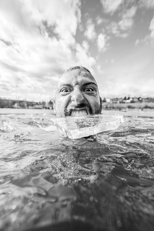 Weitwinkelaufnahme eines Mannes, der im Wasser schwimmt und ein Stück Eis zwischen den Zähnen hat. Schwarz-Weiß.