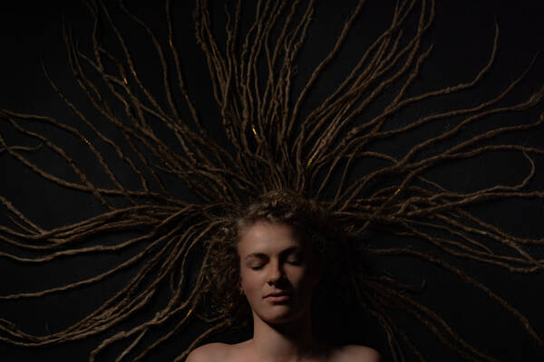 Blick von oben auf eine Frau, die auf einem dunklen Untergrund liegt, ihre langen Dreadlocks sind wie ein Strahlenkranz um ihren Kopf angeordnet.