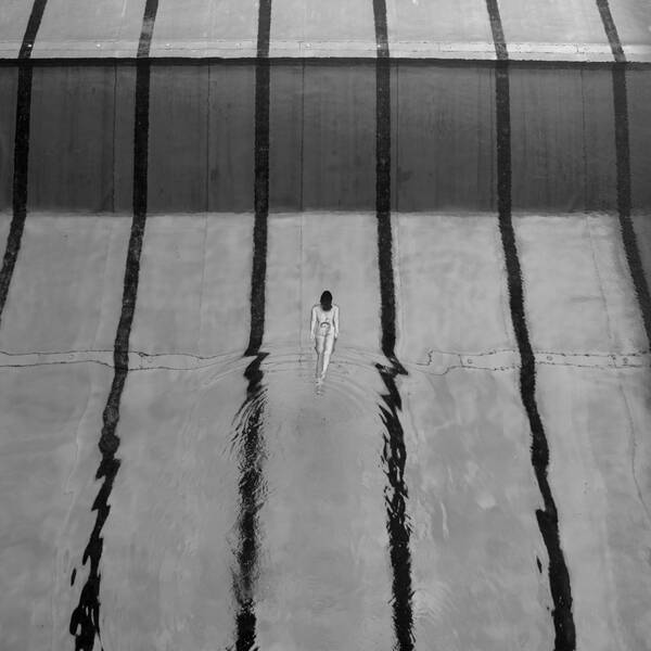 Sicht von oben auf ein Schwimmbad, im Zentrum zwischen den Bahnen schwimmt eine einzelne, nackte Frau. Schwarz-Weiß.