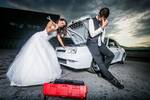 Brautpaar, die Braut beugt sich mit einem riesigen Schraubenschlüssel über den Motorraum eines weißen Sportwagens