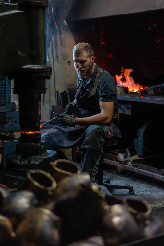 Arbeitsportrait eines jungen, bärtigen Mannes, der in einer Schmiede arbeitet: Er sitzt mit einer Schutzschürze und Handschuhen und hält einen glühenden Stab auf einen Amboss. Im Hintergrund sieht man ein Feuer in einer Esse. Im Vordergrund sind metallische Gegenstände aufgetürmt.