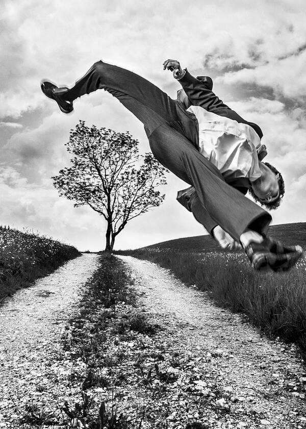 Ein Mann in Anzug macht einen extremen Luftsprung auf einem Feldweg. Schwarz-Weiß.