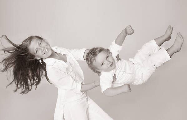 Ein Mädchen in weißem Gewand hat einen ebenfalls weiß gekleideten Buben gepackt und wirbelt ihn durch die Luft. Schwarz-Weiß.