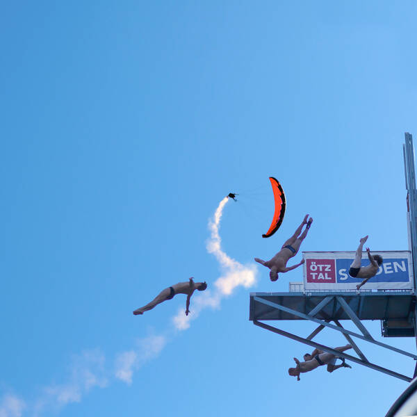 Mehrere Turmspringer vollführen gleichzeitig in unterschiedliche Richtungen Kunstsprünge von einem Sprungturm. Im Hintergrund sieht man einen Motorgesteuerten Gleitschirmflieger.