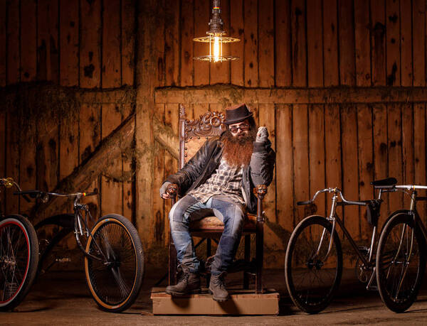 Ein Bärtiger Mann mit Hut sitzt auf einem thronartigen Sessel zwischen ausgefallenen Fahrrädern.