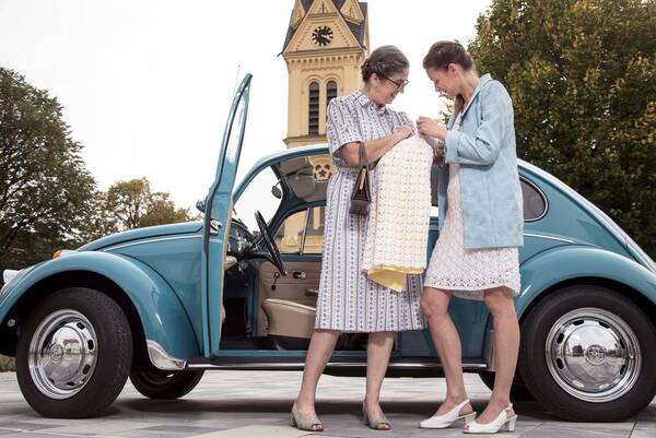 Zwei Frauen stehen vor einem blauen VW Käfer und halten ein Baby im Taufkleid. Im Hintergrund steht ein Kirchturm. Vintage-Look.