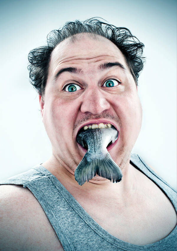 Ein Mann in grauem Trägershirt hat einen ganzen Fisch im Mund, so dass nur die Schwanzflosse herausschaut.