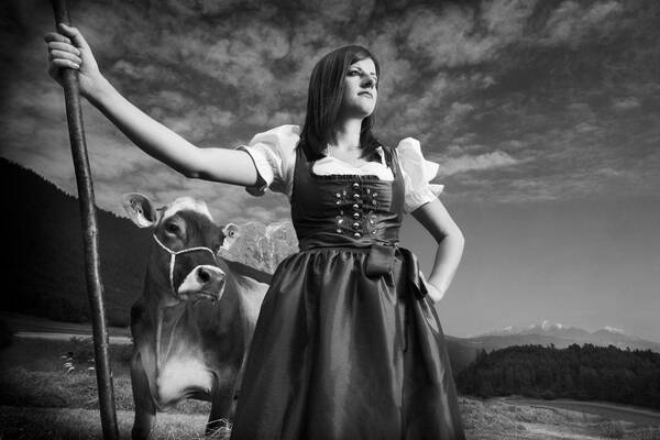 Eine junge Frau in einem Trachenkleid steht in heroischer Pose mit einem Haselnuss-Stecken. Hinter ihr steht eine Kuh. Schwarz-Weiß.