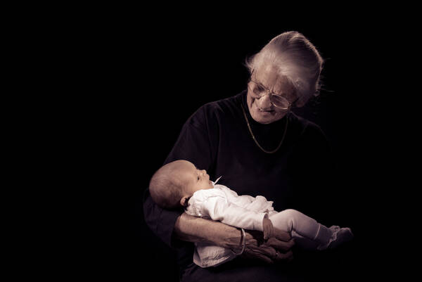 Eine dunkel gekleidete, brillentragende alte Frau mit einem Goldkettchen hält ein weiß gekleidetes Baby im Arm.