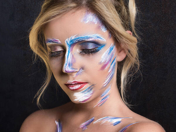 Portrait einer jungen, blonden Frau, deren Gesicht und Schulterbereich bemalt ist.