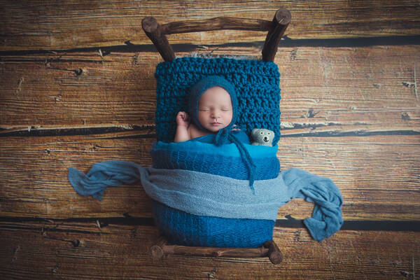 Ein Neugeborenes liegt mit einer blauen Strickhaube in einem blau gehäkelten rustikalen Bettchen mit blauen Decken und einem kleinen Teddybären. Blick von oben.