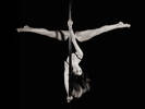 Eine Frau in dunklem Trikot hängt kopfüber an einer Pole-Dance-Stange und macht mit den Beinen einen Spagat. Schwarz-Weiß.