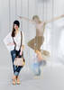 Eine Frau in bunten Highheels, Jeans mit Hosenträgern und weißer Oversize-Bluse trägt eine schwarze Mütze und eine elegante Damenhandtasche. hinter ihr hängen Ketten von der Decke, hinter denen teiltransparent und bewegungsunscharf ein Mann mit nacktem Oberkörper Tanzt und ein weiterer zu Boden fällt.