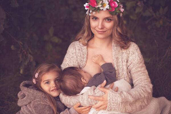 Eine Frau mit Blumenkrone stillt in der Natur ein Kind. ein zweites Kind schmiegt sich dazu.