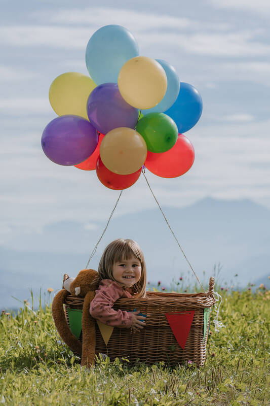 Ein kleines Mädchen sitzt mit einem Kuscheltier in einem großen Korb, an dem viele bunte Luftballons befestigt sind. 