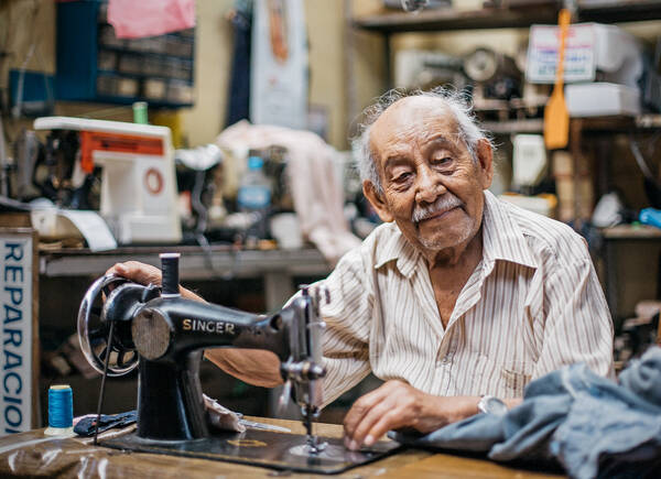 Ein alter Mann sitzt im gestreiften Hemd an einer alten Singer-Nähmaschine und lächelt in die Kamera.