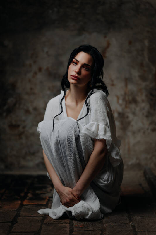 Eine Frau sitzt mit traurigem Gesichtsausdruck auf einem groben Klinkerboden und hat ihre Knie umschlungen. Von der Wand im Hintergrund blättert die Farbe ab.