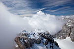 Verschneite Berglandschaft, im Hintergrund Nebelbänke unter blauem Himmel. Ganz klein hebt sich die Silhouette von vier Menschen ab, die durch den Schnee zum Abstieg gehen.