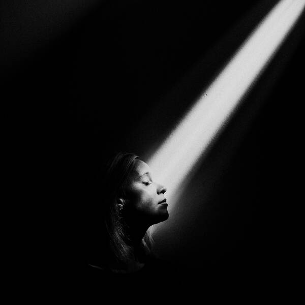 Portrait einer jungen Frau im Profil. Ein Lichtstrahl fällt durch die Dunkelheit auf ihr Gesicht. Schwarz-Weiß. Low key.