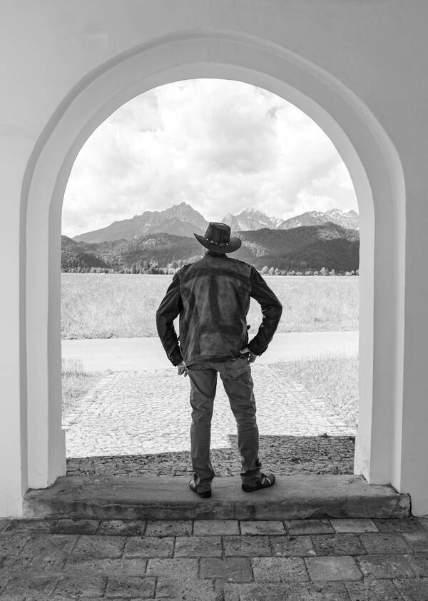 Ein Mann mit Cowboyhut  steht in einem Torbogen und blickt in die bergige Ferne. Schwarz-Weiß