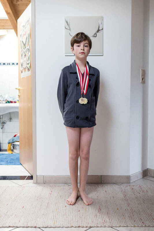Ein Bub ohne hose aber mit einem zweireihigen grauen Jackett hat zwei Medaillien umgehängt und steht so vor einem Bild, dass das dort abgebildete Hirschgeweih aus dem Kopf des Buben zu kommen scheinen.