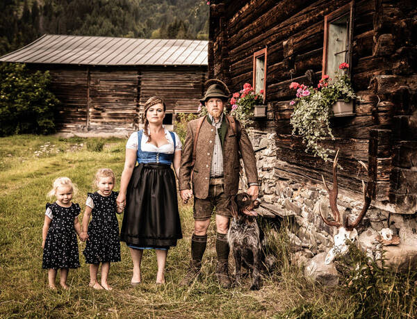 Familienportrait eines Jägers, seiner Frau und zweier Mädchen mit dem Jagdhund vor Holz-Blockhäusern. Im Vordergund liegt eine Hirsch-Trophäe.