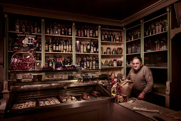 Blick in einen Greissler-Laden mit Spirituosen in den Regalen und Süßspeisen in den Vitrinen. Der Verkäufer steht hinter dem Tresen.